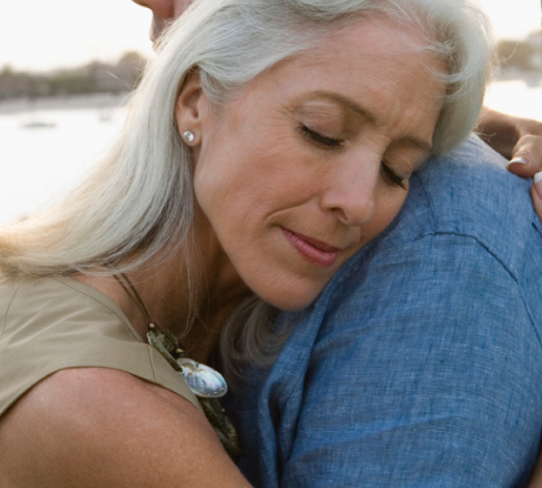 Woman with gray hair hugs man in denim shirt with lake in background.