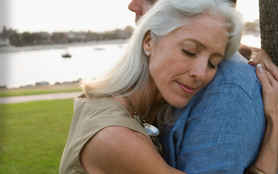 Woman with gray hair hugs man in denim shirt with lake in background.
