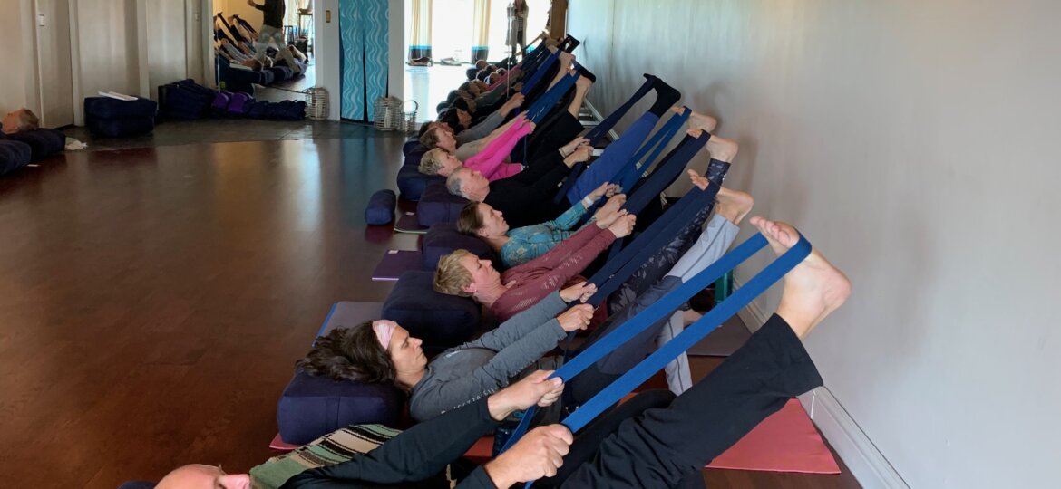 Mixed age class demonstrates Avita Yoga lying supine with strap around right foot and leg extended in the air.