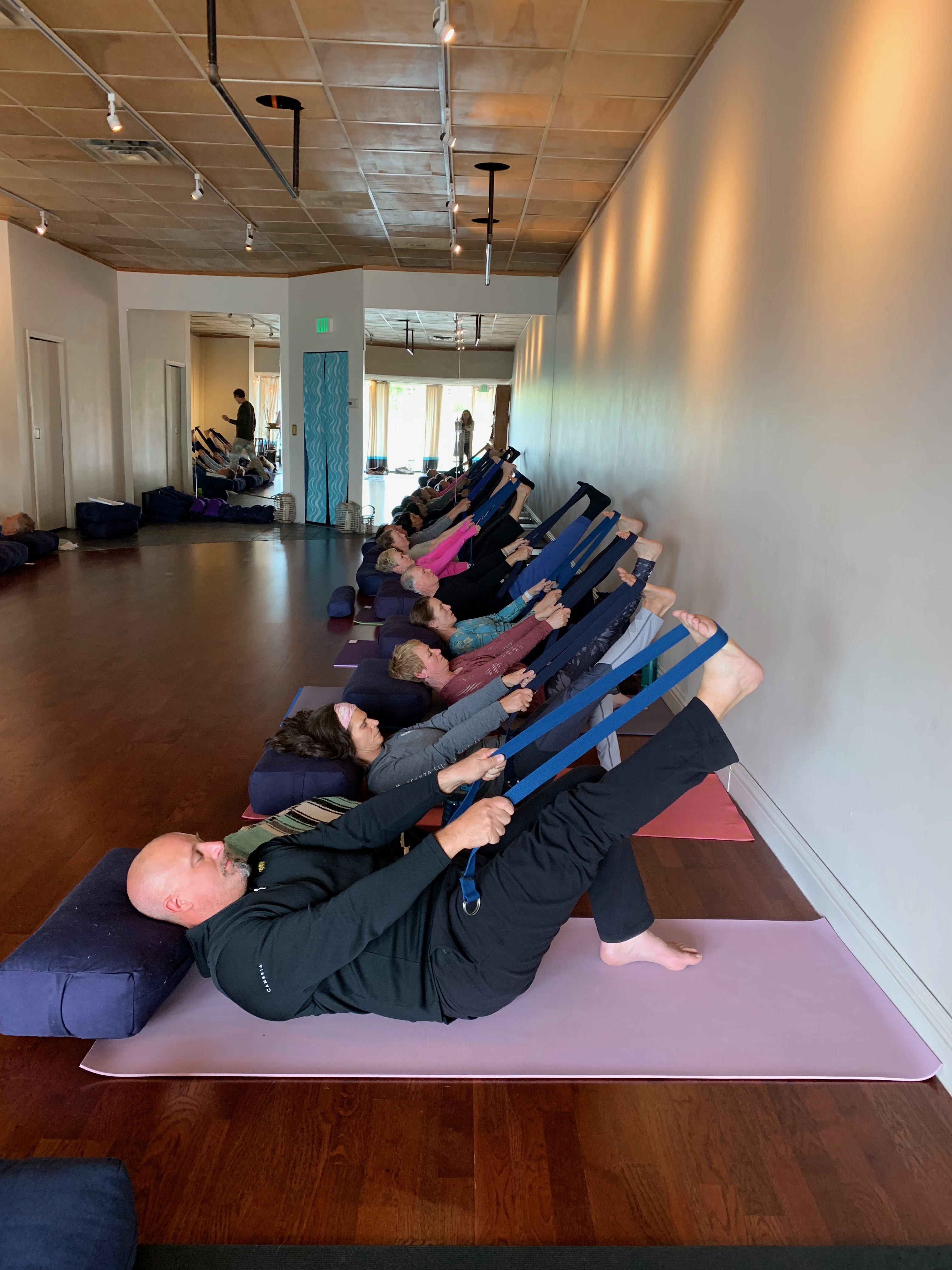 Mixed age class demonstrates Avita Yoga lying supine with strap around right foot and leg extended in the air.